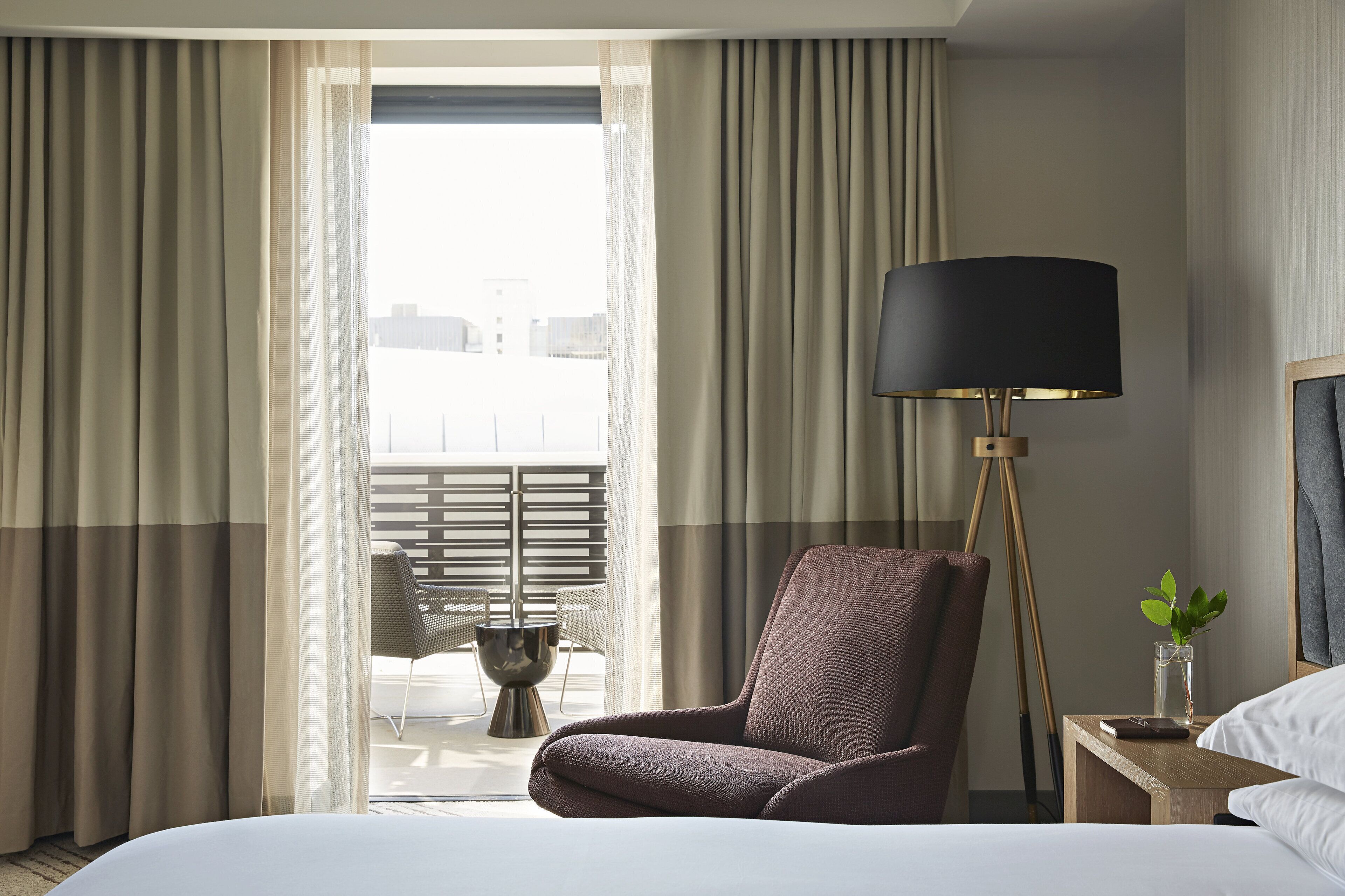 A hotel room with a bed, a chair, a nightstand with a lamp, and a view of the outdoors through the window.