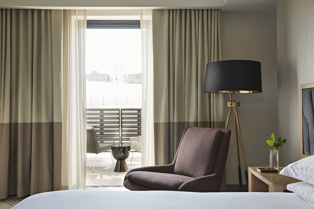 A hotel room with a bed, a chair, a nightstand with a lamp, and a view of the outdoors through the window.