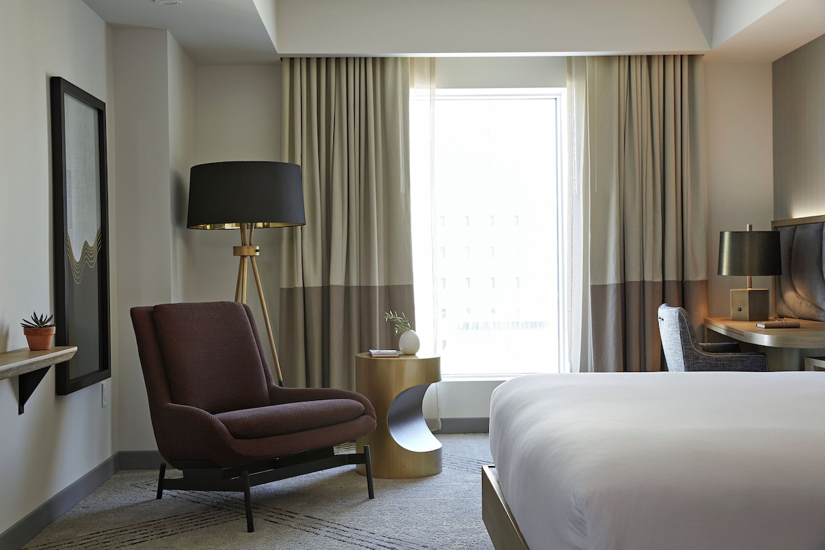 A hotel room with a bed, a desk, a chair, a lamp, and a TV.