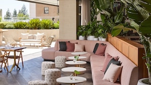 A modern outdoor seating area with a sofa, ottomans, and round tables, surrounded by large potted plants and a view of the city. - Kimpton Sawyer Hotel by IHG (Sacramento)