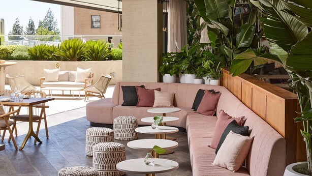 Un espacio moderno al aire libre con sofá, pufs y mesas redondas, rodeado de grandes macetas con plantas y vistas a la ciudad.