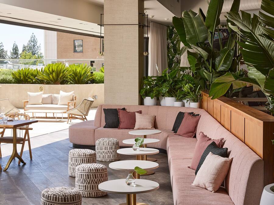 A modern outdoor seating area with a sofa, ottomans, and round tables, surrounded by large potted plants and a view of the city.