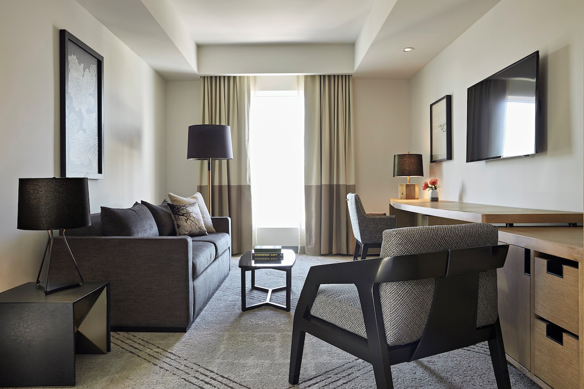A modern hotel room with a sofa, armchair, desk, and a flat-screen TV.