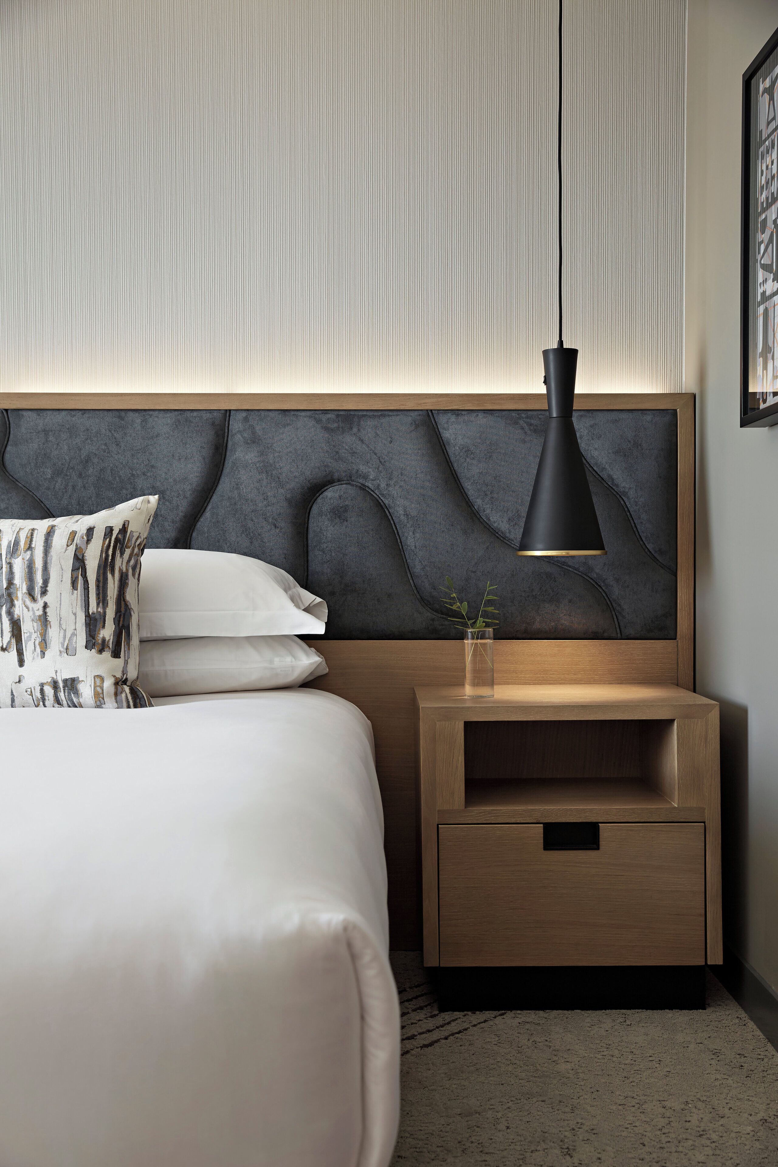 Un dormitorio moderno con una cama, una mesita de noche con lámpara y una pared con textura.
