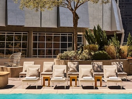 Zona de piscina con sillones de descanso y una pequeña mesa, rodeada de follaje y la fachada moderna de un edificio.