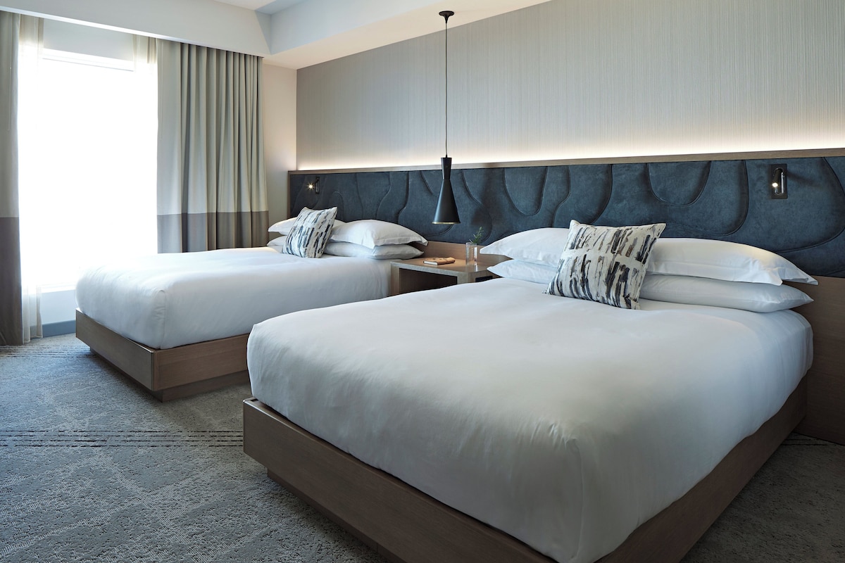 A hotel room with two beds, a nightstand, a lamp, and a large window with curtains.