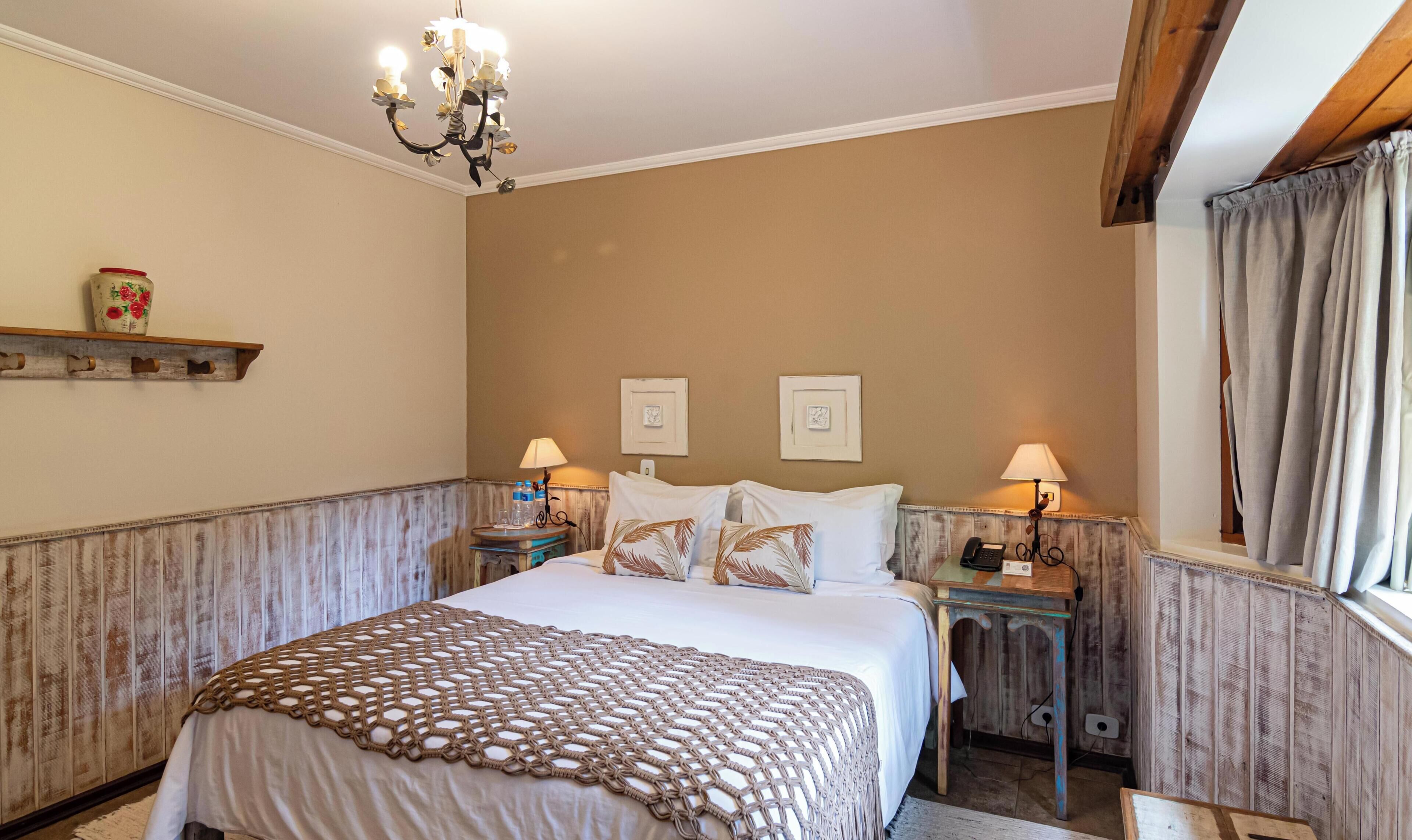A bedroom with a large bed, two bedside tables, a chandelier, a wooden headboard, and a window with curtains.
