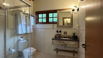 A bathroom with a glass shower enclosure, a toilet, a sink with a mirror above it, and a window with a view of greenery.