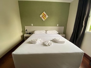Una habitación de hotel con cama, almohadas, una mesita de noche, una obra de arte en la pared y una ventana con cortinas.