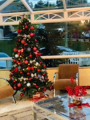 Un sapin de Noël décoré d’ornements et de guirlandes lumineuses, une table basse en verre agrémentée de décorations rouges et un fauteuil marron dans une pièce dotée d’un plafond en verre.