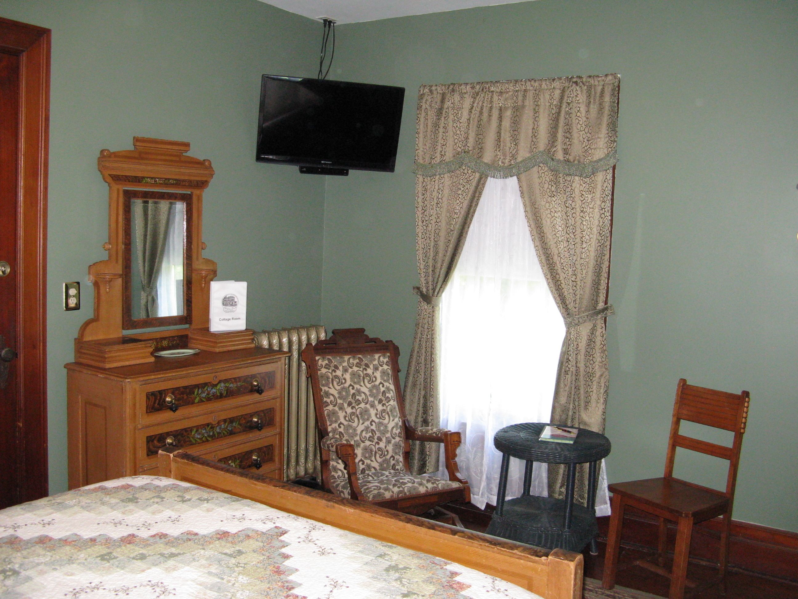A bedroom with a wooden bed, dresser, and chairs, a television mounted on the wall, and a window with curtains.