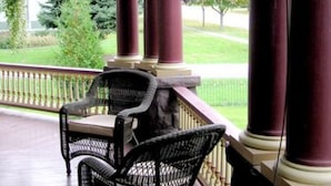 A covered porch with two wicker chairs and a table, supported by columns with a hanging flower basket. - 1897 Beekman House (Dundee)