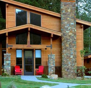 A modern log cabin with a stone chimney, large windows, and a front porch with red chairs.