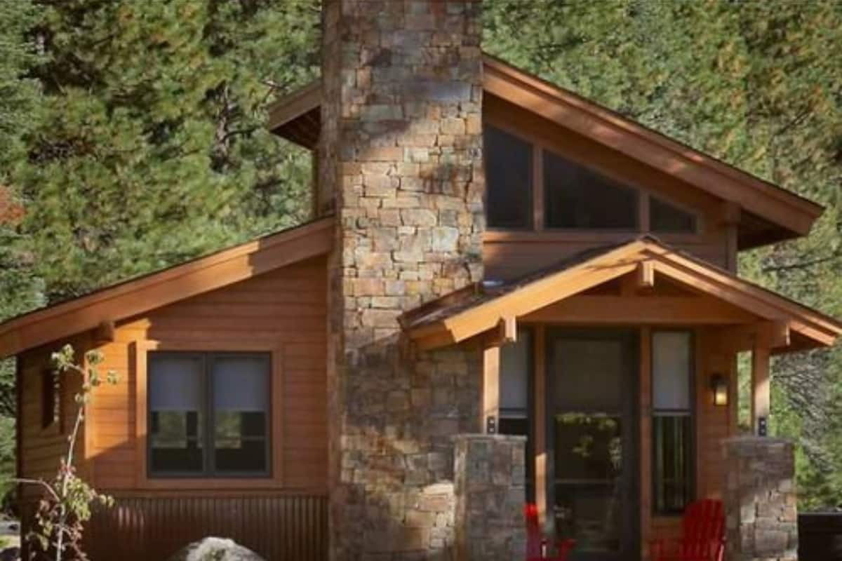 Una cabaña de troncos moderna con chimenea de piedra, ventanas amplias y un porche cubierto con sillas rojas.