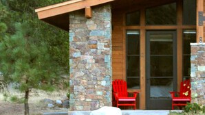 A modern house with a stone exterior, large glass doors, and two red Adirondack chairs in front.