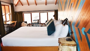 Un grand lit avec du linge de lit blanc, une tête de lit en bois et une lampe de chevet. On aperçoit, au fond, une petite table avec une lampe et une chaise.