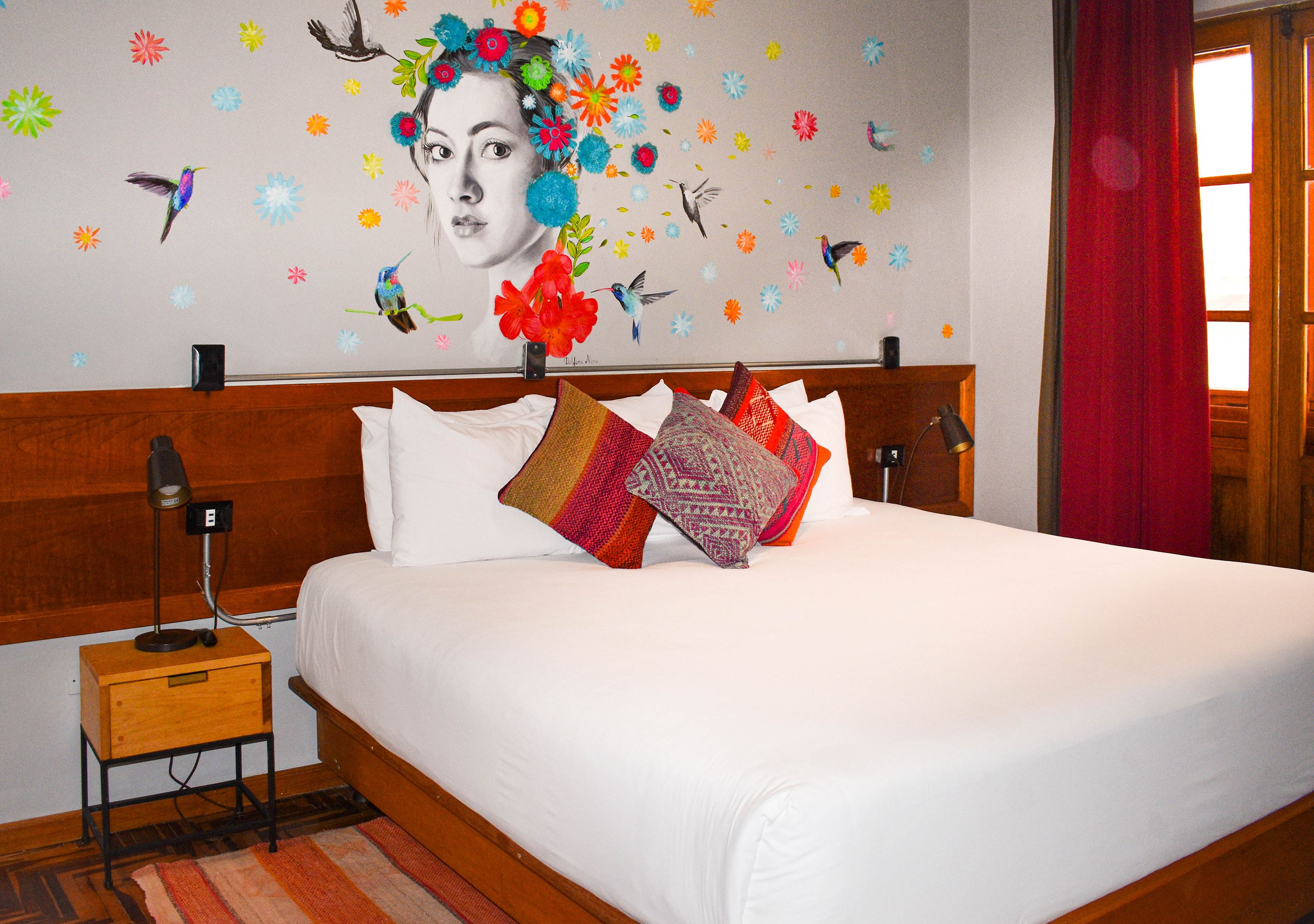 A hotel room with a large bed, a nightstand, a lamp, and a wall with colorful floral and bird decals.