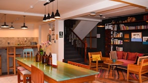 Un vaste espace de vie comprenant une table à manger en bois, plusieurs chaises, un canapé et une bibliothèque remplie de livres. On aperçoit un escalier au fond de la pièce, menant à l’étage supérieur.