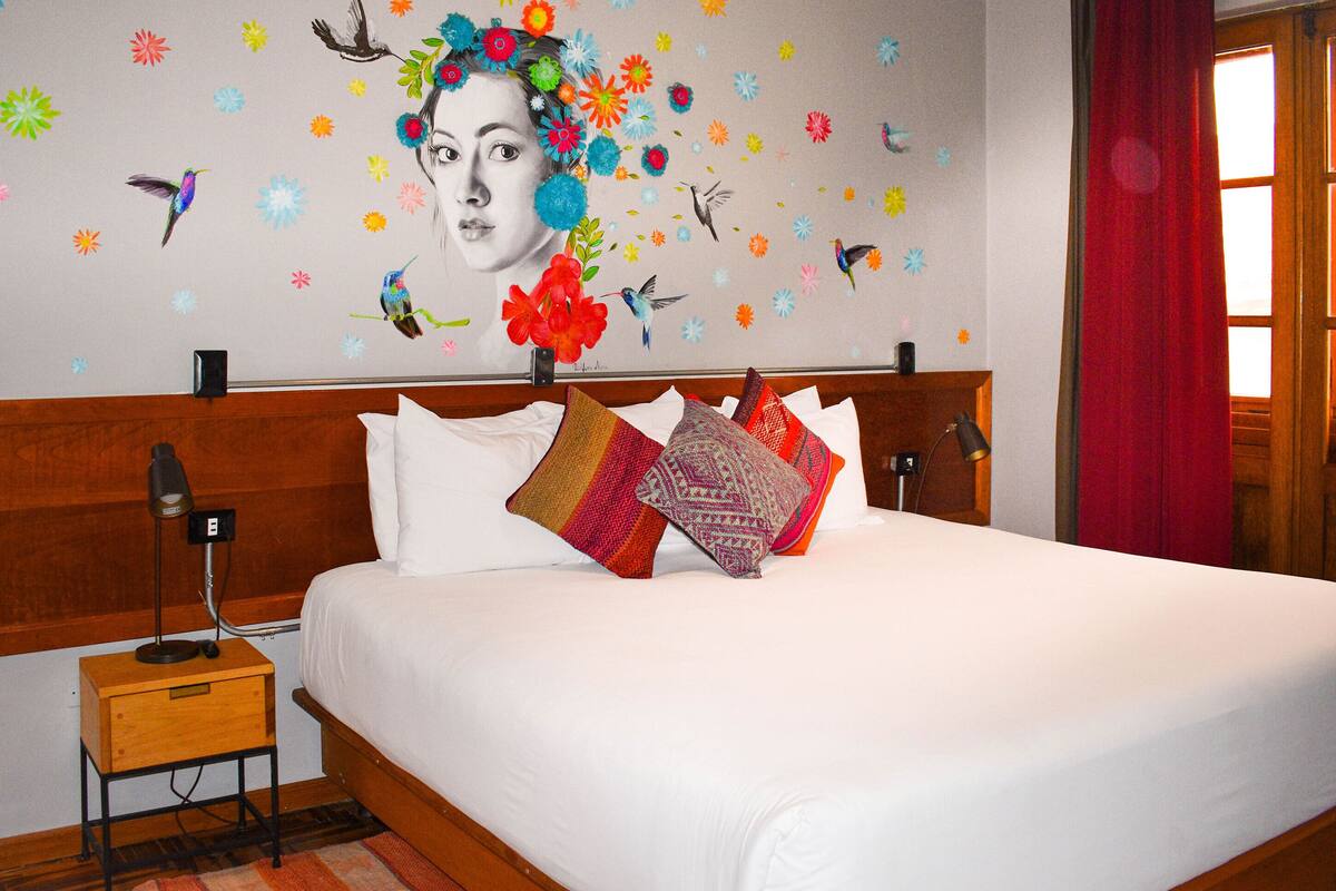 Una cama bien hecha con sábanas blancas y almohadas coloridas, un cabecero de madera, una mesita de noche con una lámpara y una pared decorada con adhesivos de flores y pájaros.