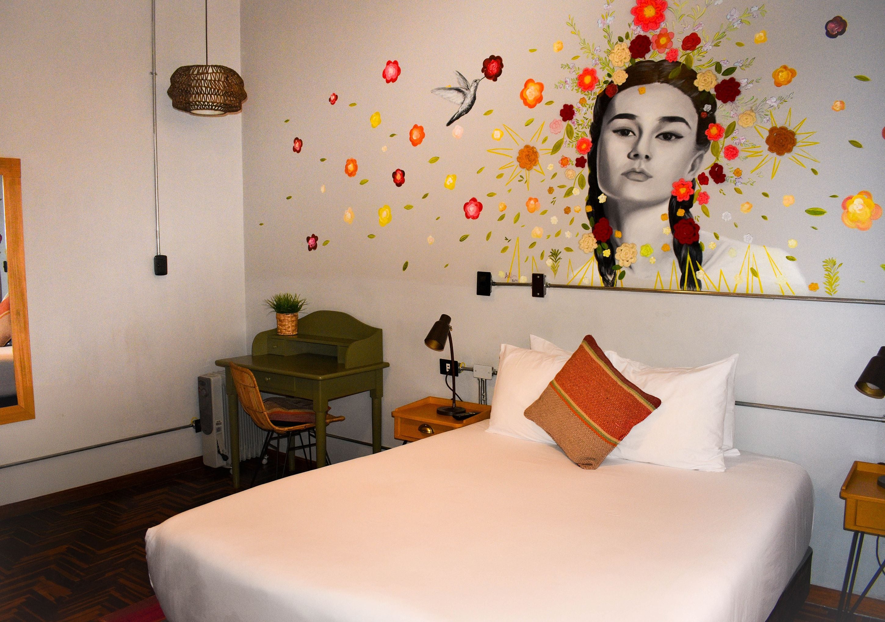 A hotel room with a bed, a desk, a chair, a lamp, and a wall with floral and bird decorations.