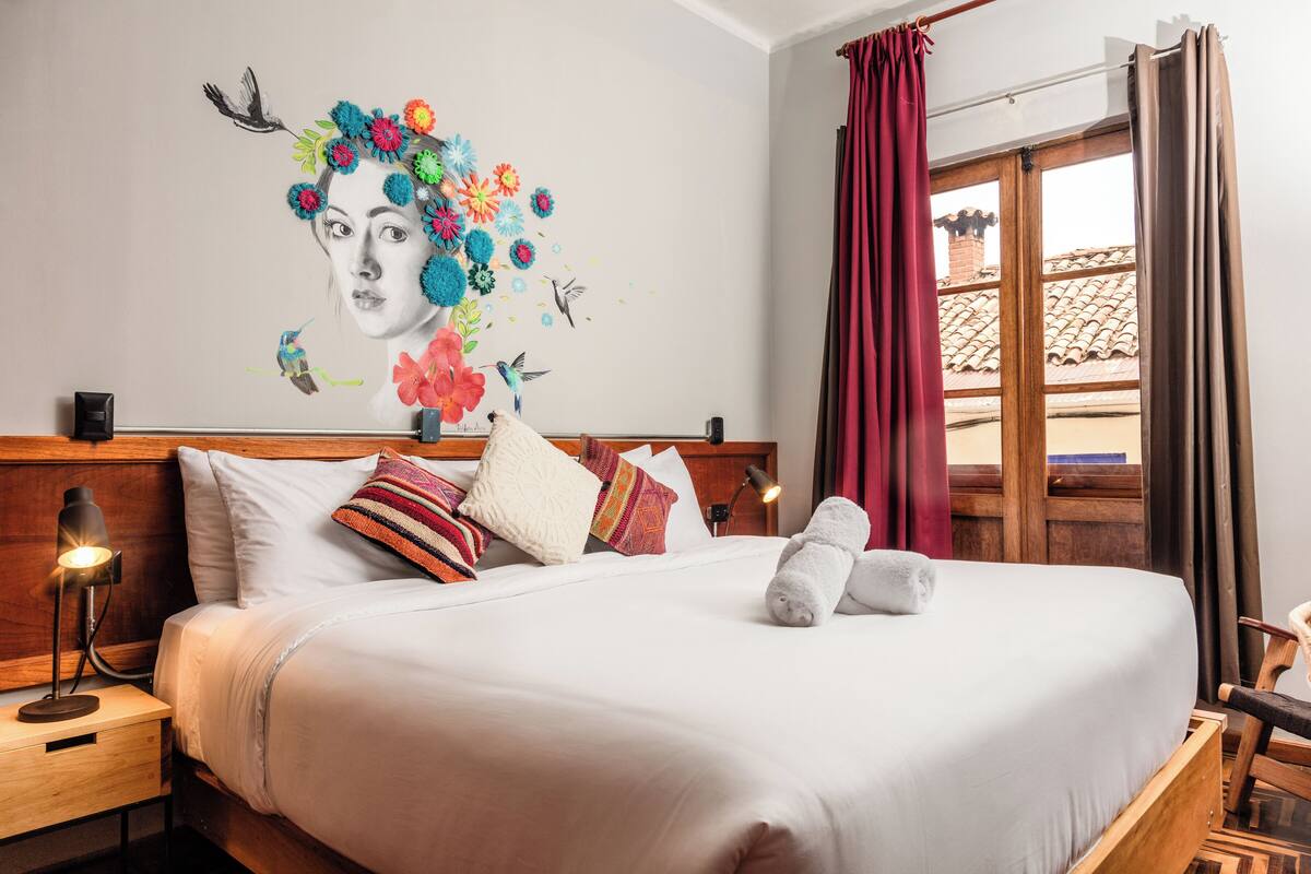 Una cama bien tendida con sábanas blancas, cabecera de madera y almohadones decorativos. Un par de toallas está colocadas sobre la cama.