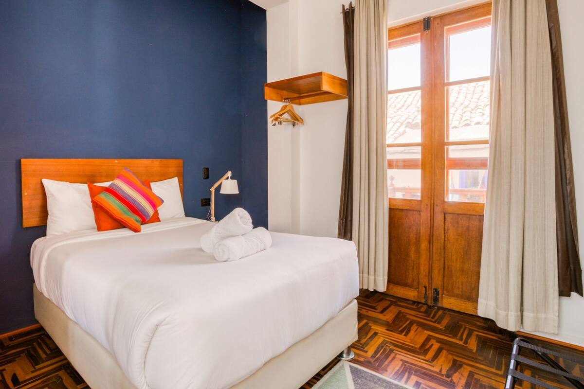 Habitación de hotel con cama, cabecera de madera, lámpara colgante, estante de madera y vista al exterior a través de una puerta.