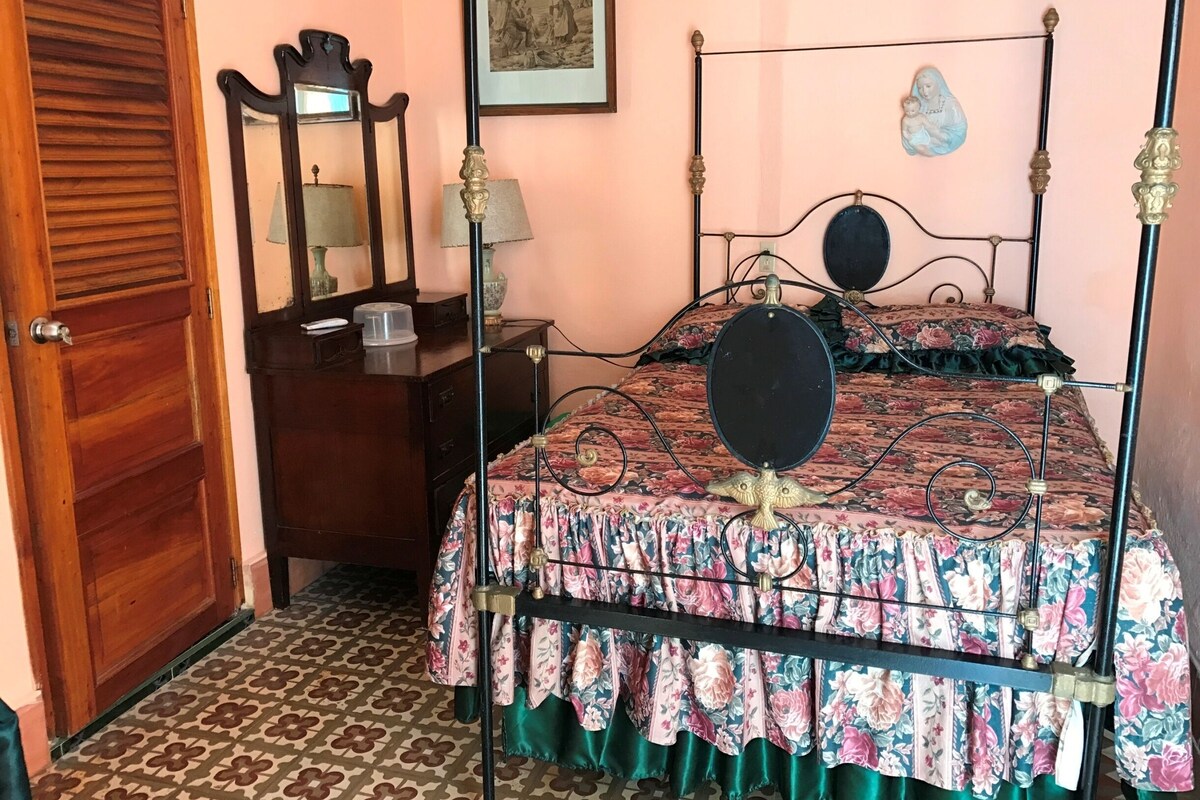 Un dormitorio con cubrecama floral, una puerta de madera, un tocador con espejo y un cuadro en la pared.