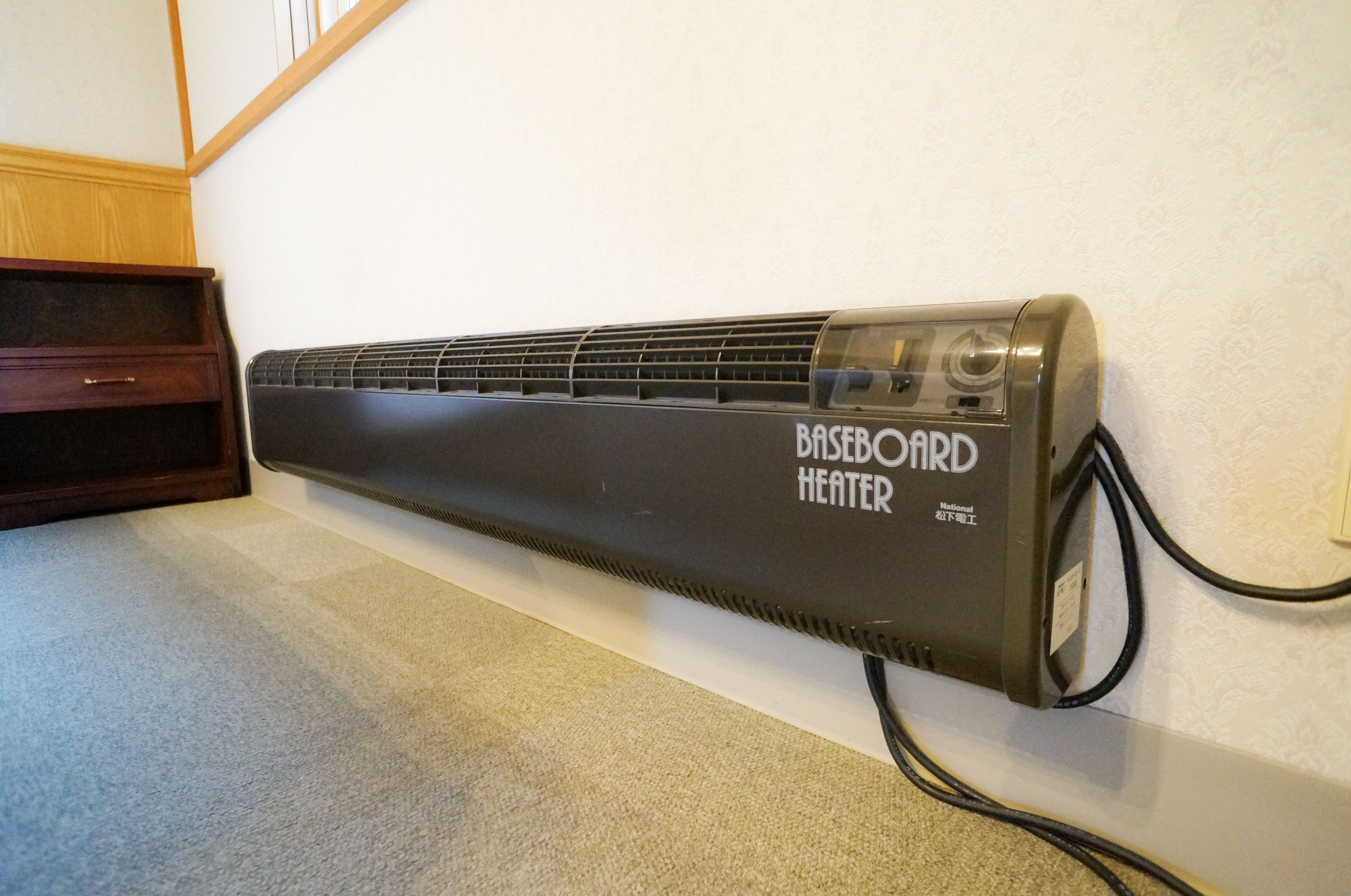 A baseboard heater mounted on a wall in a room with a carpeted floor and a wooden cabinet.