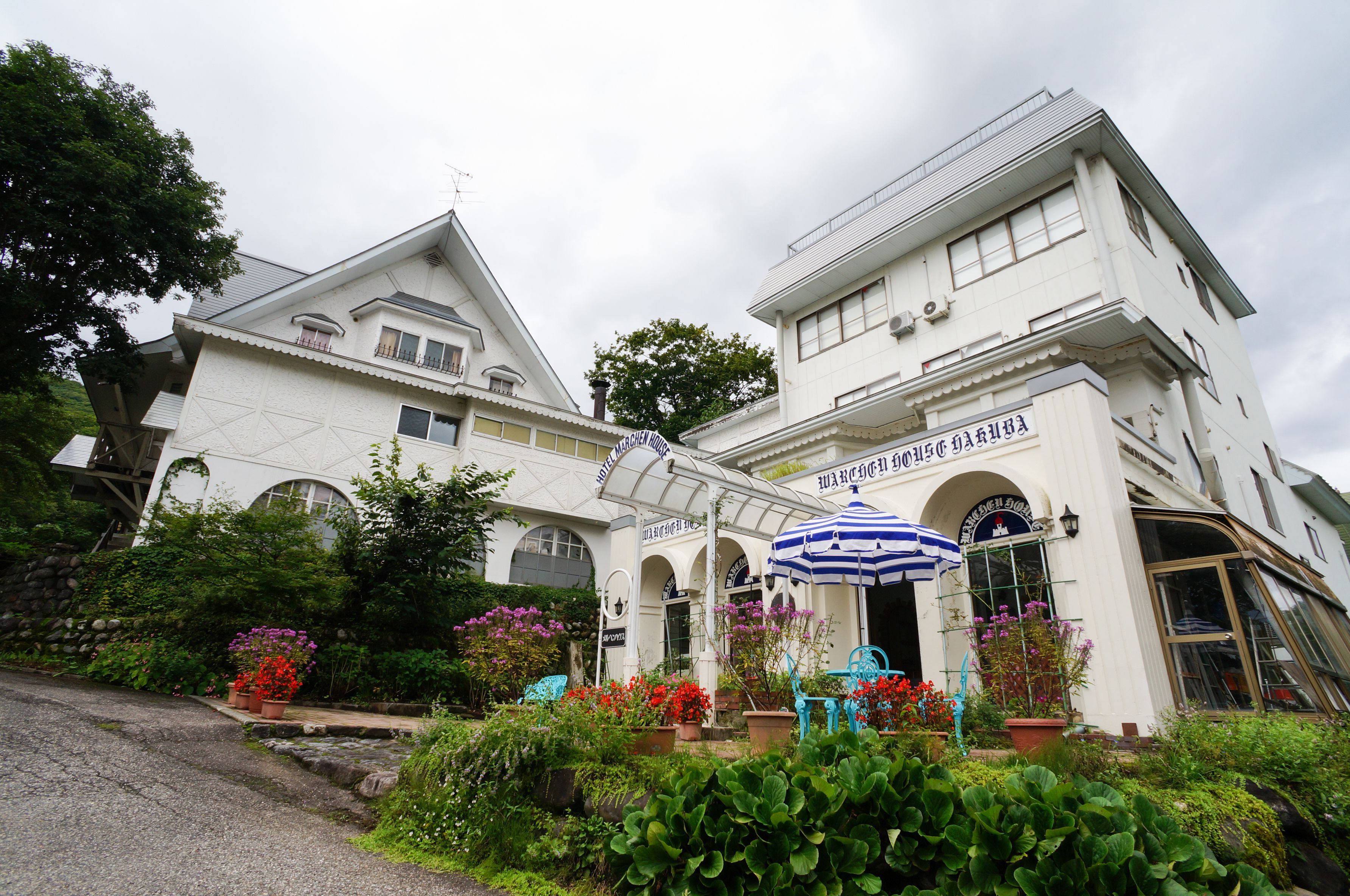 A white building with a sign that reads "Hotel". There are potted plants and a blue umbrella in front of the building.