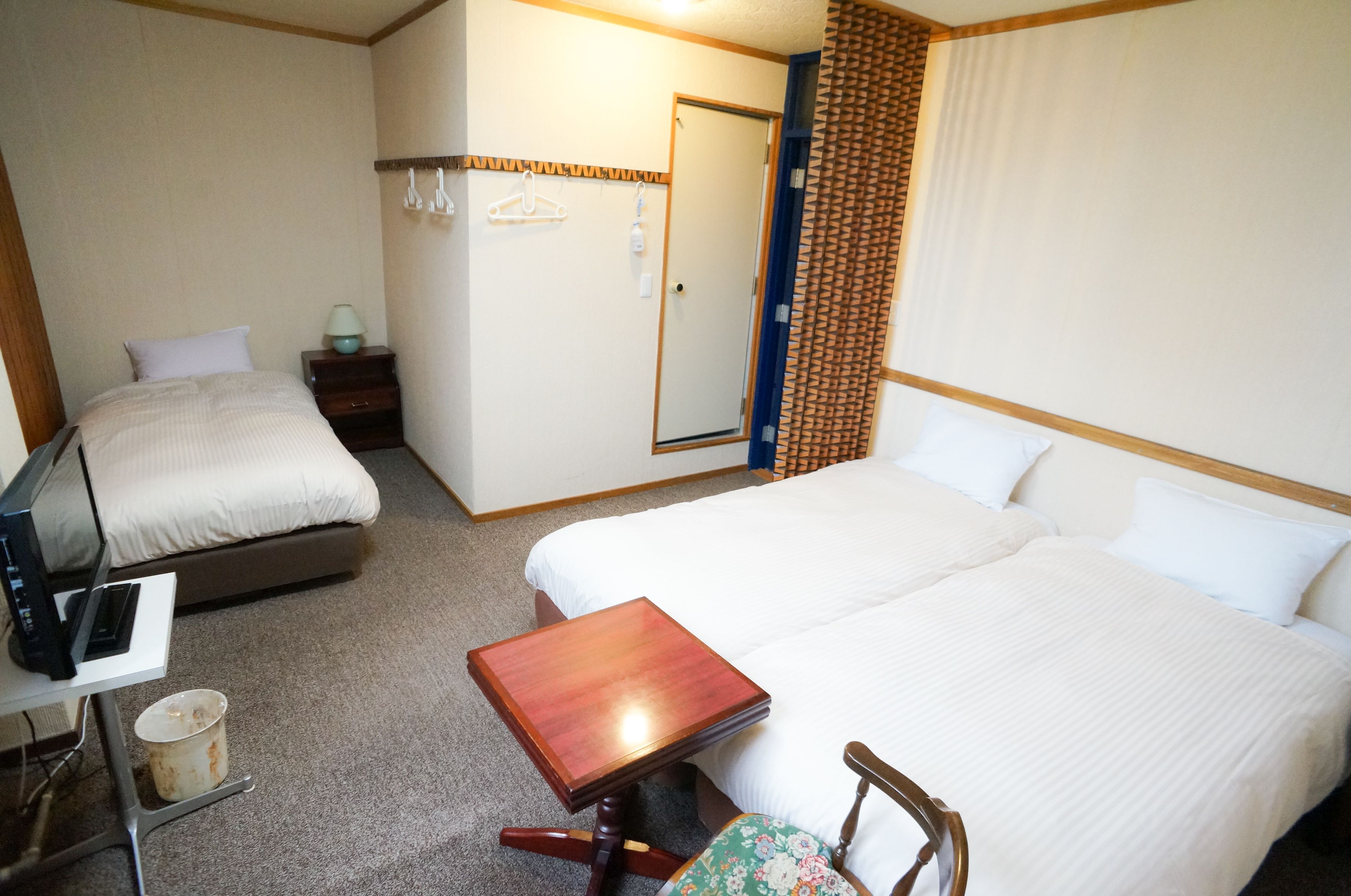 A hotel room with two beds, a small table, a chair, a desk with a computer, and a trash bin.