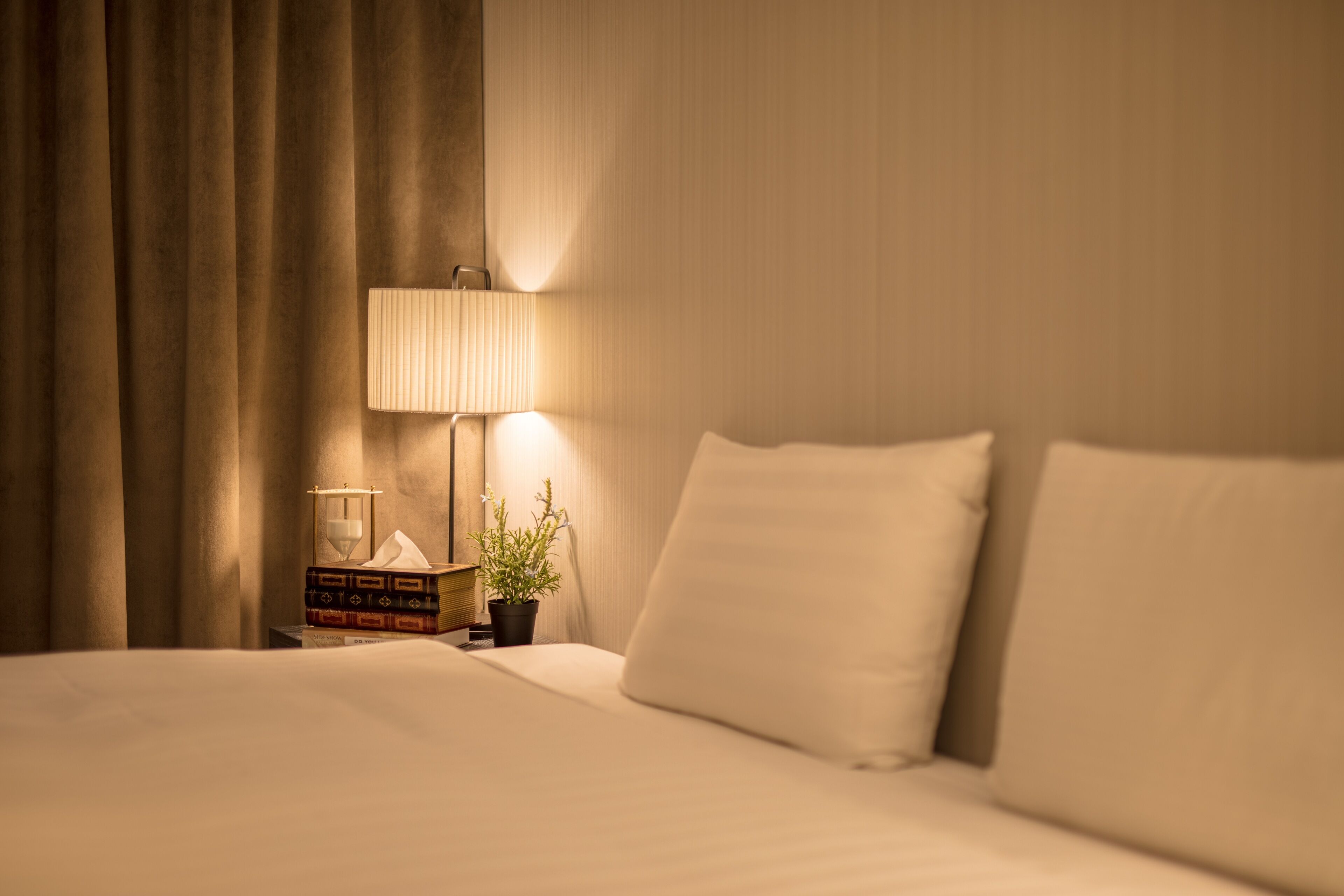 Una cama bien tendida con sábanas blancas, una lámpara de noche, una pequeña planta en maceta y una pila de libros en la mesita de noche.