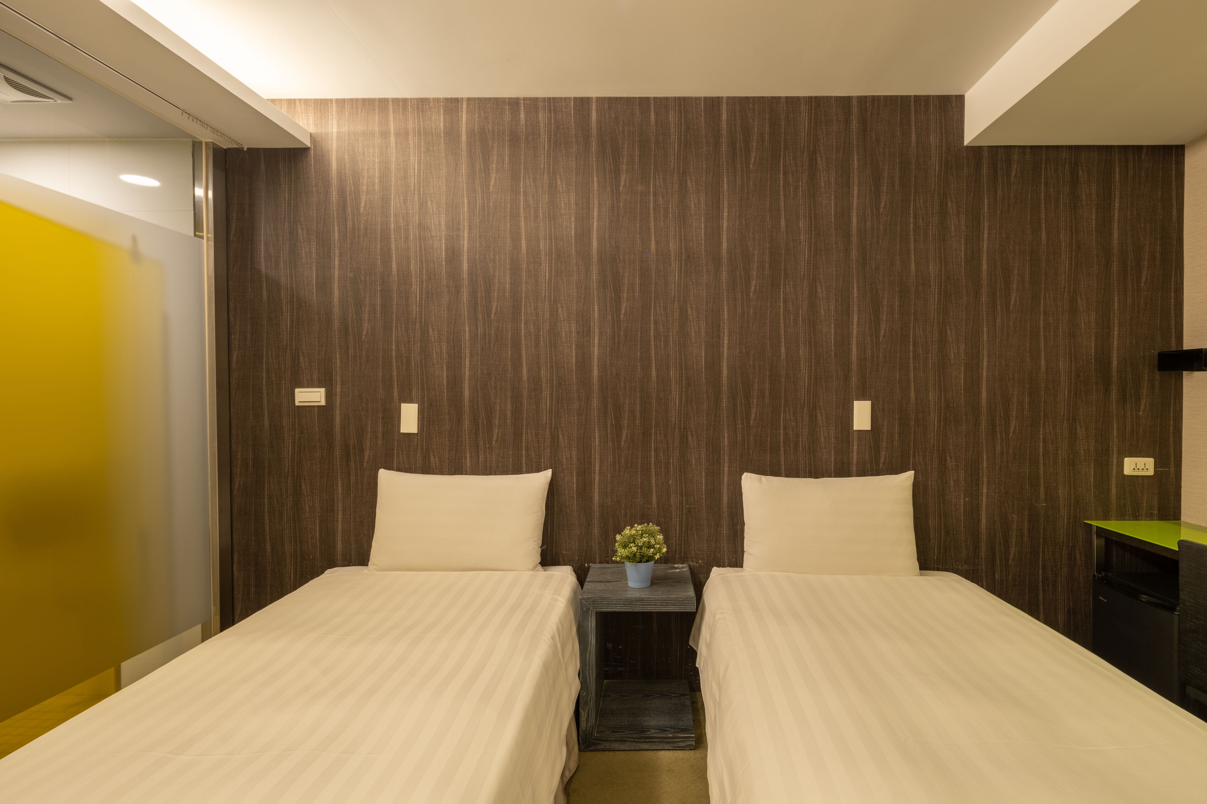 A hotel room with two beds, a small table with a plant, and a green desk.