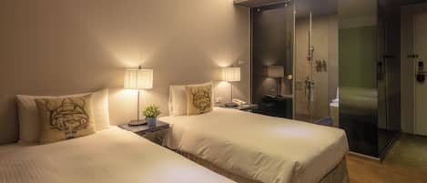 Quarto de hotel com duas camas, luminárias de cabeceira, uma mesinha com uma planta e área de chuveiro em vidro.