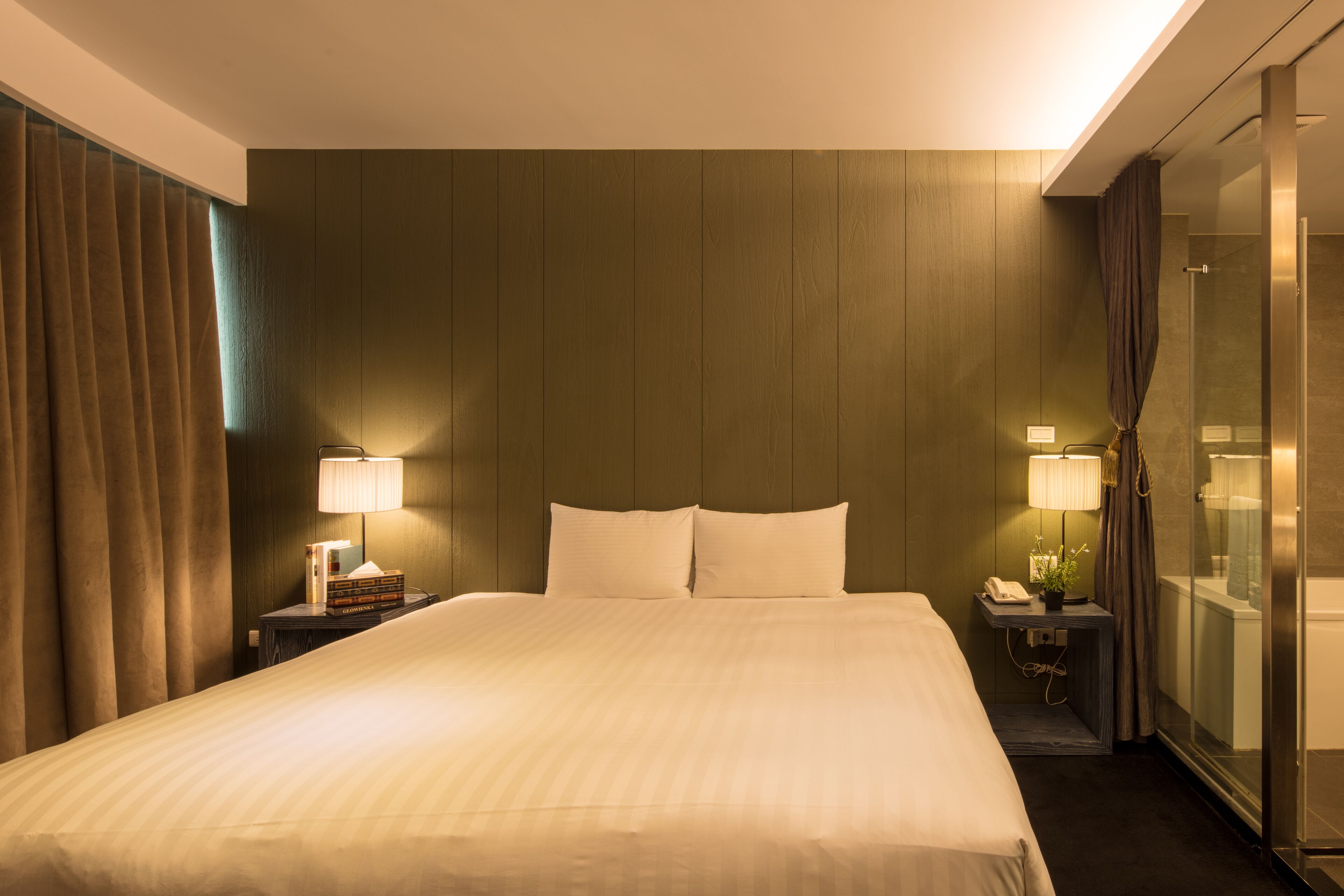 A hotel room with a large bed, two bedside tables with lamps, a nightstand with a phone and a plant, and a glass shower enclosure.