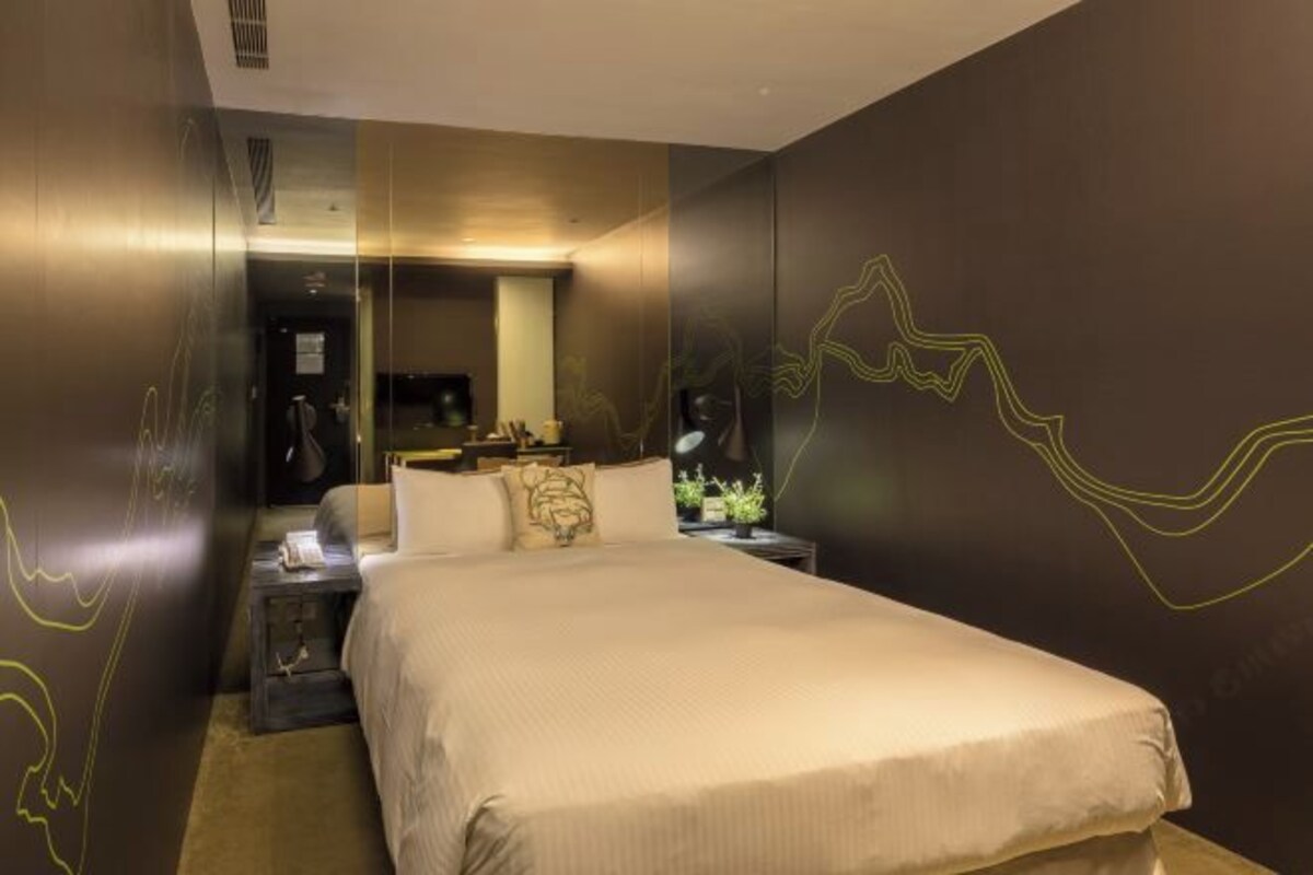 A hotel room with a large bed, a bedside table, a nightstand, a mirror, and a wall with abstract line art.