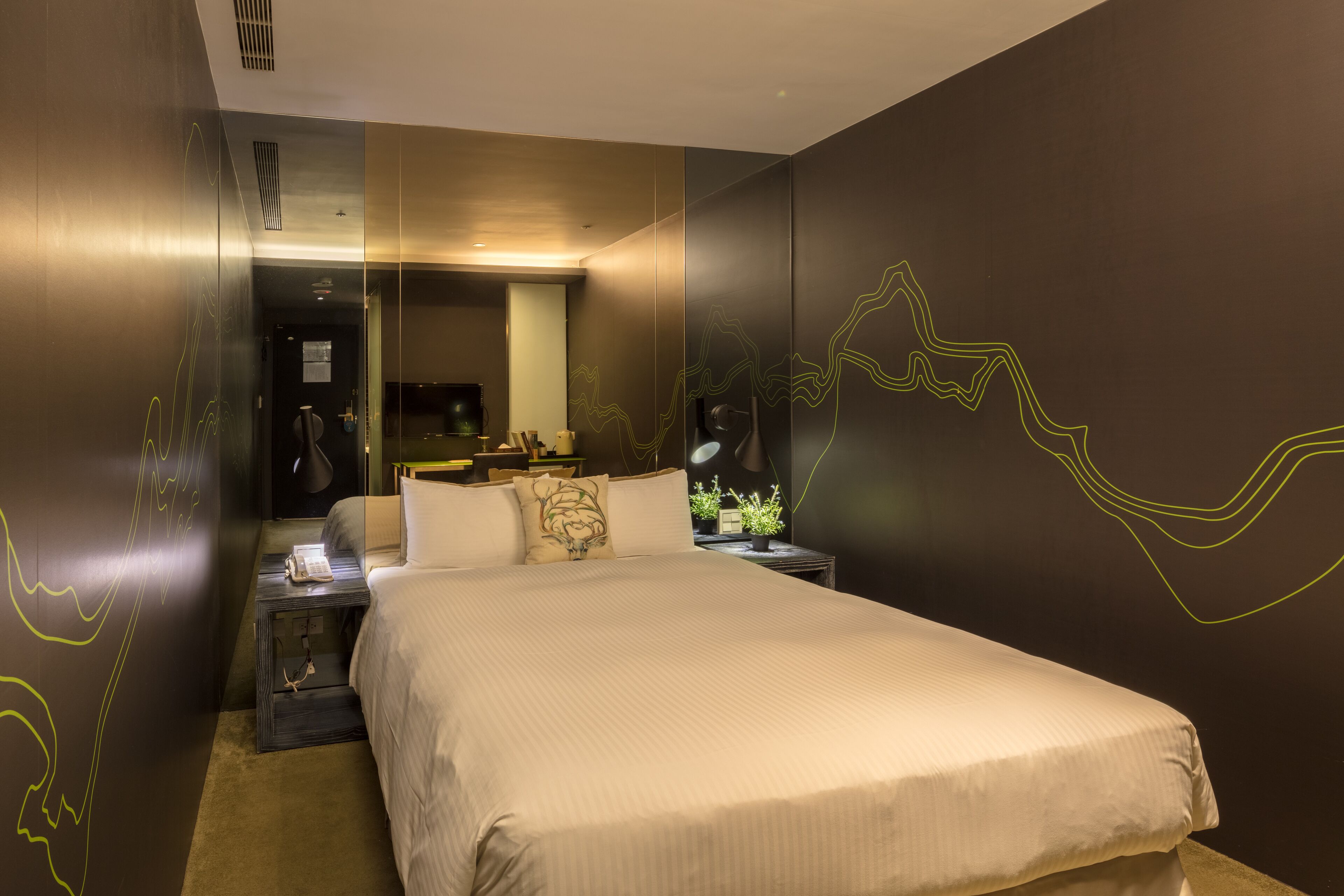 A hotel room with a large bed, a bedside table with a lamp, a mirror, and a wall with abstract line art.