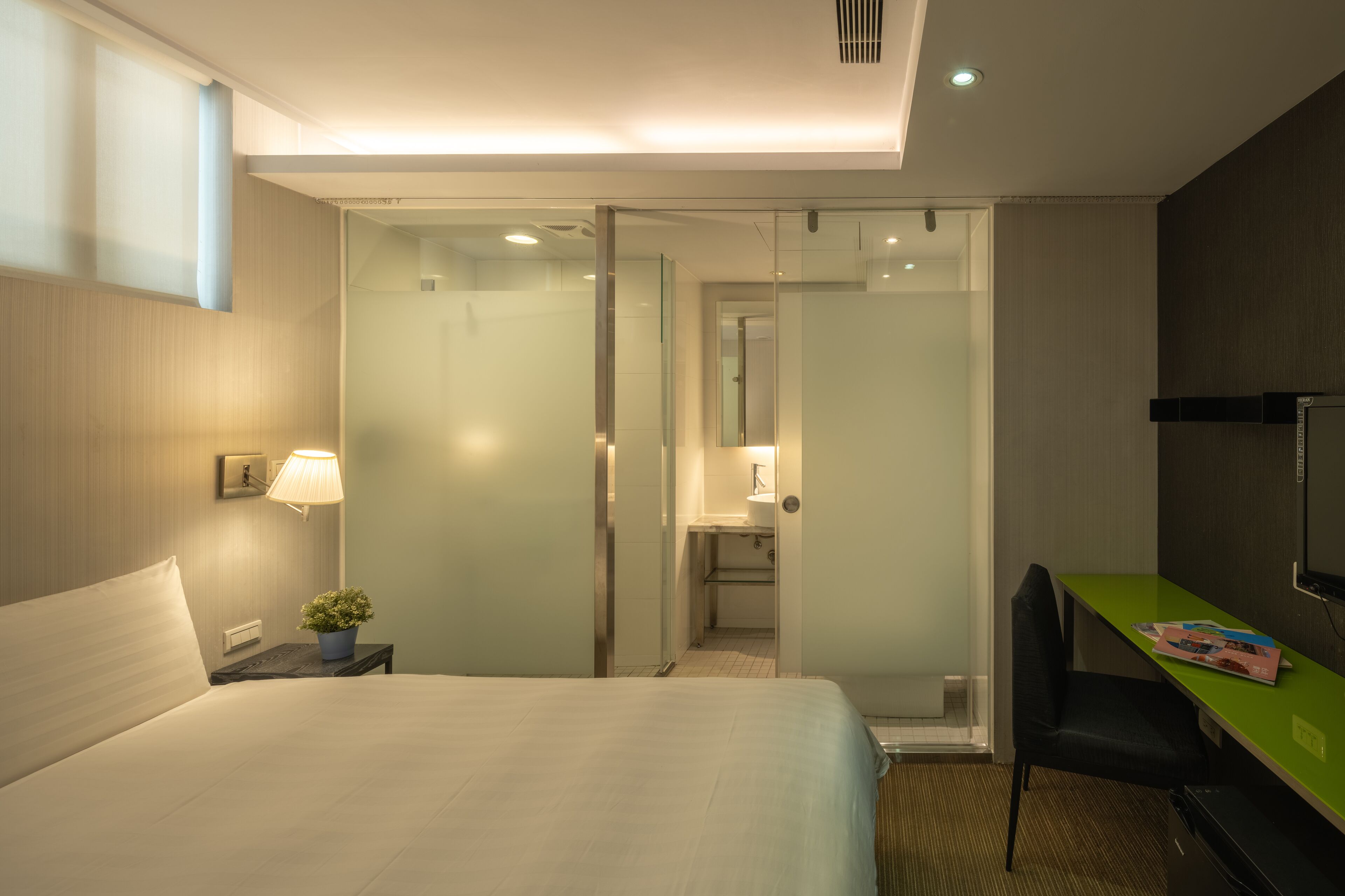 A hotel room with a bed, a desk, a chair, a lamp, and a small plant.