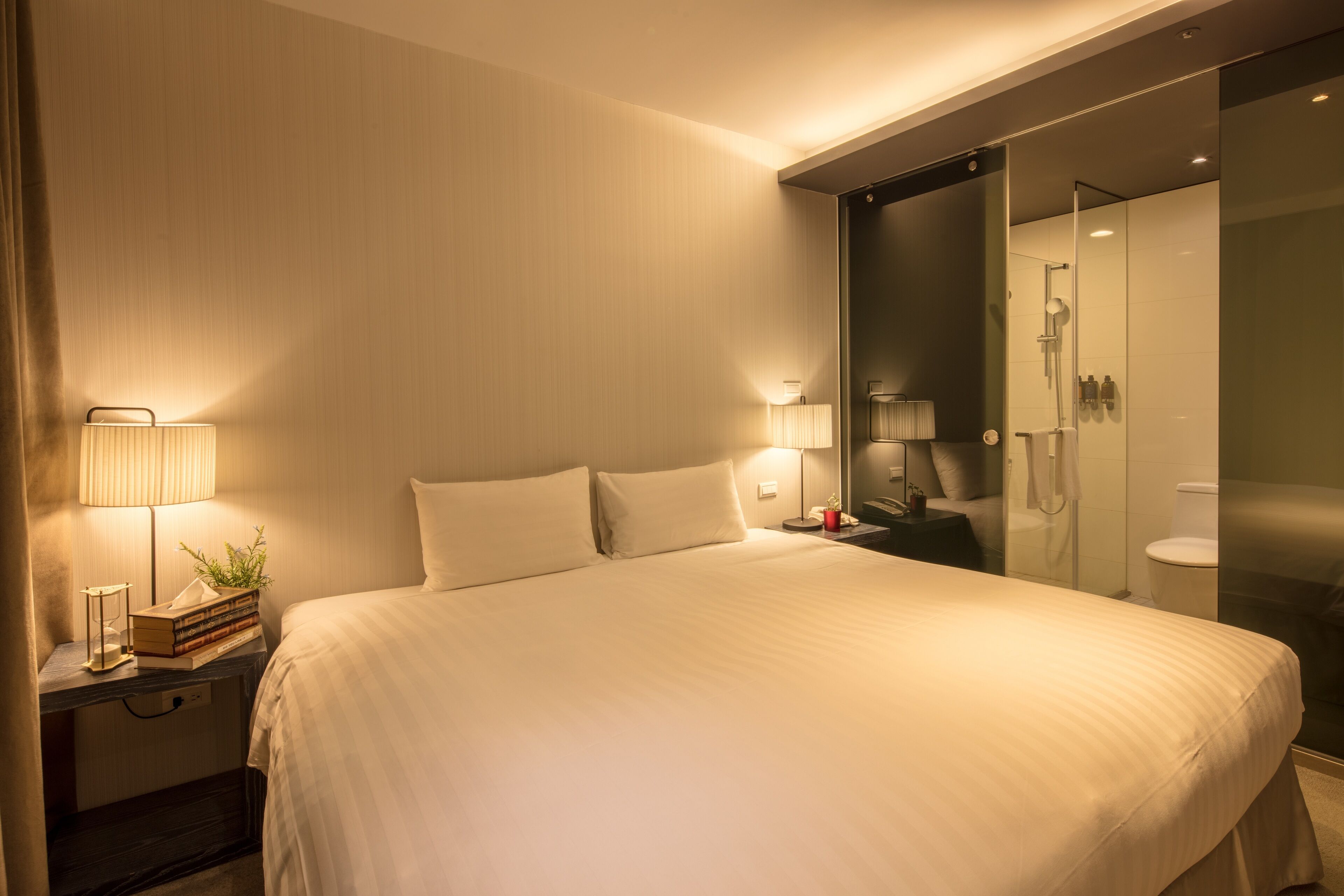 Ein modernes Hotelzimmer mit einem großen Bett, Nachttischlampen, einem Nachttisch mit Lampe und Büchern sowie einer gläsernen Duschkabine.