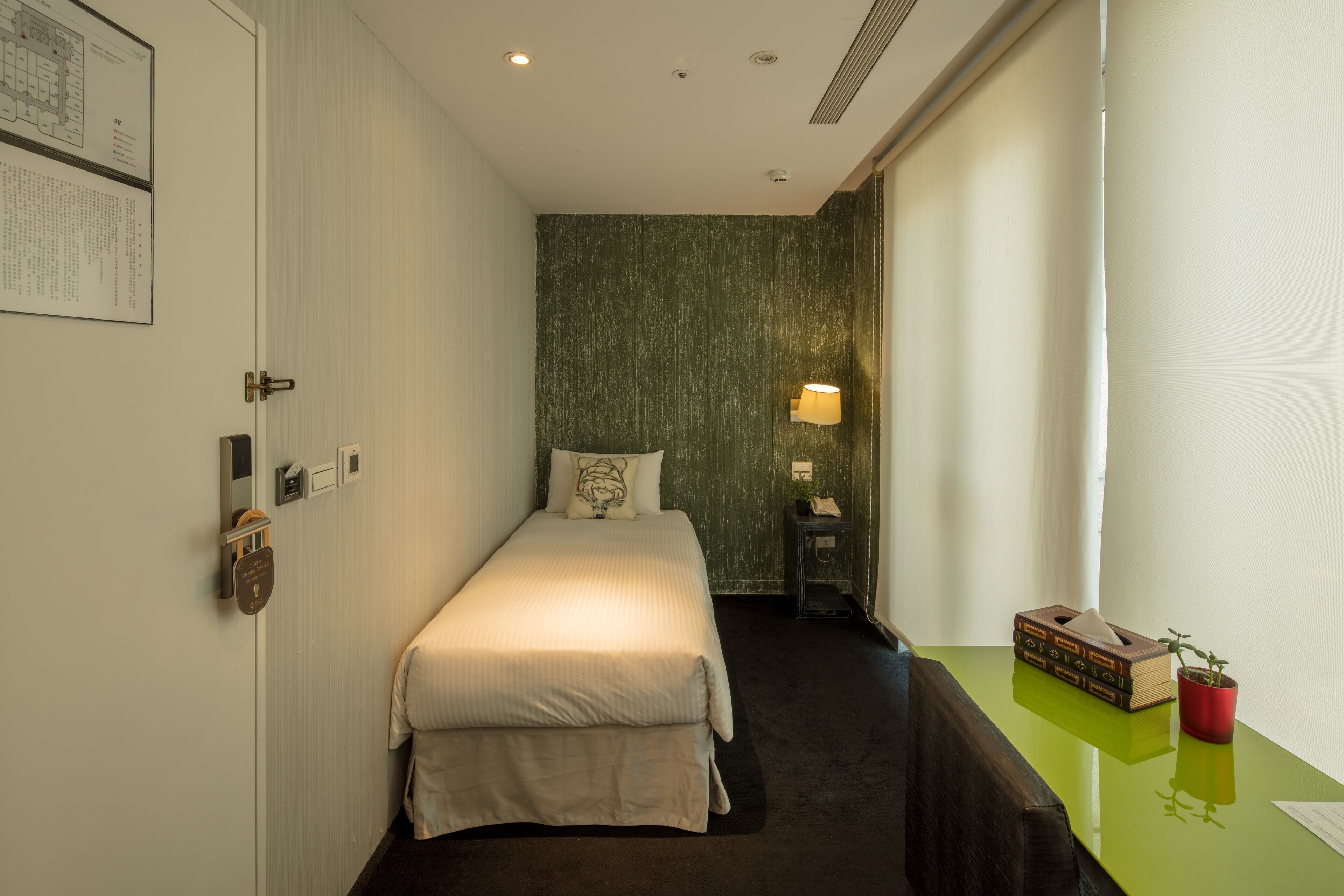 Une chambre d’hôtel étroite, avec un lit, une table de chevet, une lampe et une petite table sur laquelle se trouvent une plante et des livres.
