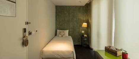 Une chambre d’hôtel étroite, avec un lit, une table de chevet, une lampe et une petite table sur laquelle se trouvent une plante et des livres.