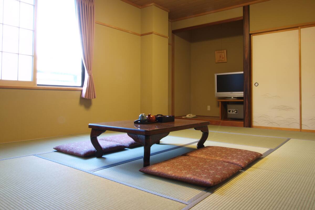 A traditional Japanese room with tatami flooring, a low table, cushions, a television, and sliding doors.
