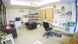 A modern living room with a sofa, a desk, and a TV. There are shelves with books and decorative items. - Hiran Residence (Patong)