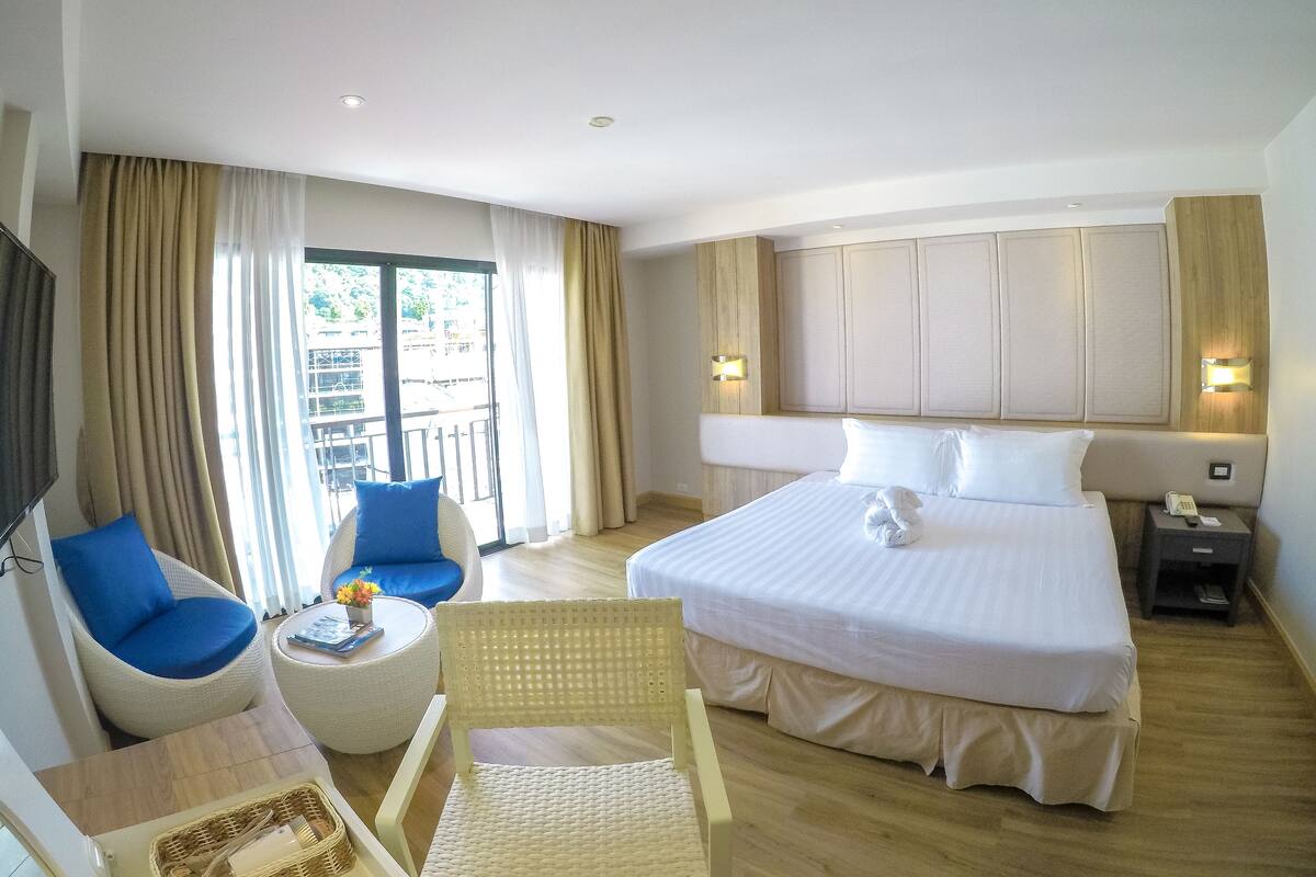 Ein Hotelzimmer mit einem großen Bett, zwei Sesseln, einem kleinen Tisch, einem Fernseher und einem Balkon mit Vorhängen.