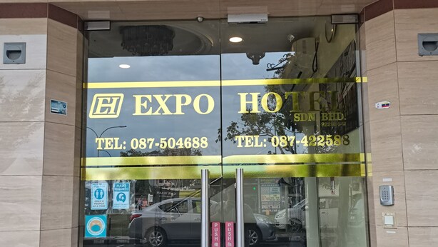 Der Eingang zum Expo Hotel mit Glastüren, einer digitalen Anzeige oberhalb des Eingangs und einem Schild mit dem Hotelnamen und der Telefonnummer.