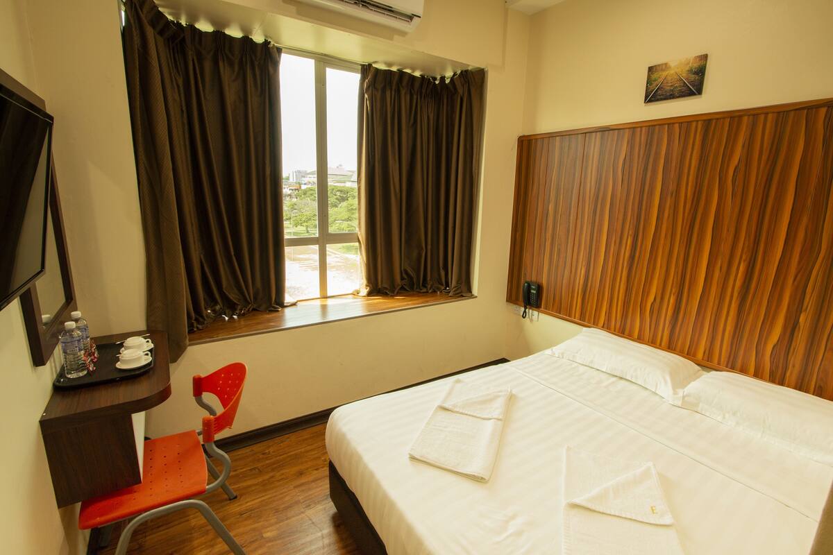 A hotel room with a bed, a desk with a chair, a window with curtains, and a wooden headboard.