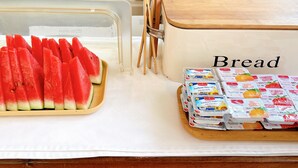 A kitchen counter with a clear plastic container holding sliced watermelon, a bread box labeled "Bread," and a tray of packaged spreads. - Check-in Resort Koh Larn (Koh Lan)