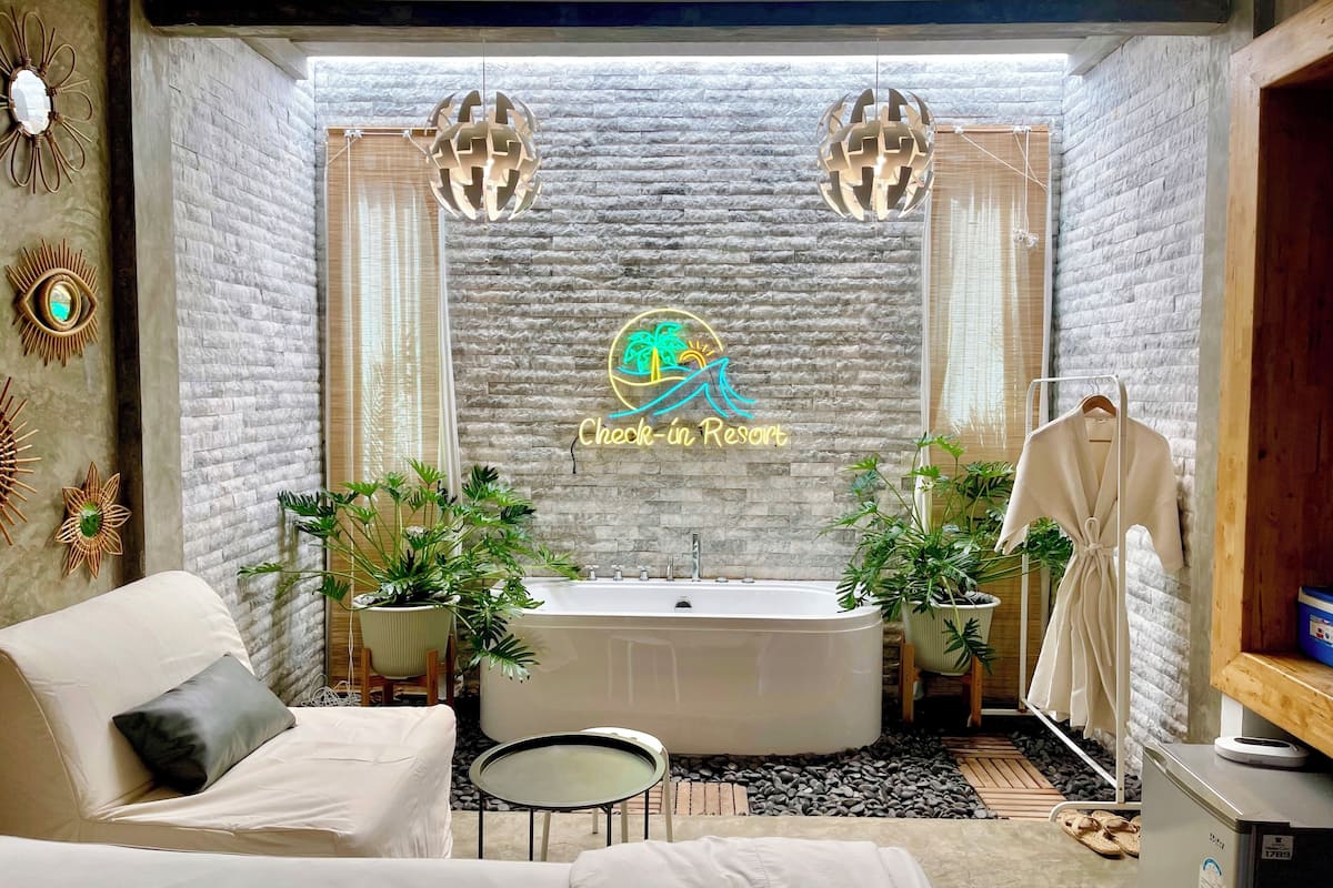 A spa room with a stone wall, a bathtub, two potted plants, a towel rack, and a small table.