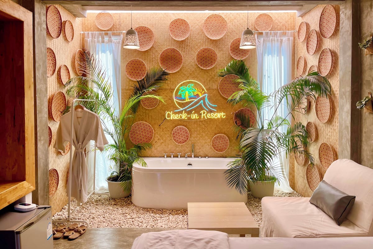 A modern bathroom with a large bathtub, a sink, and a towel rack. There are several potted plants and a neon sign that reads "Check-in Resort".