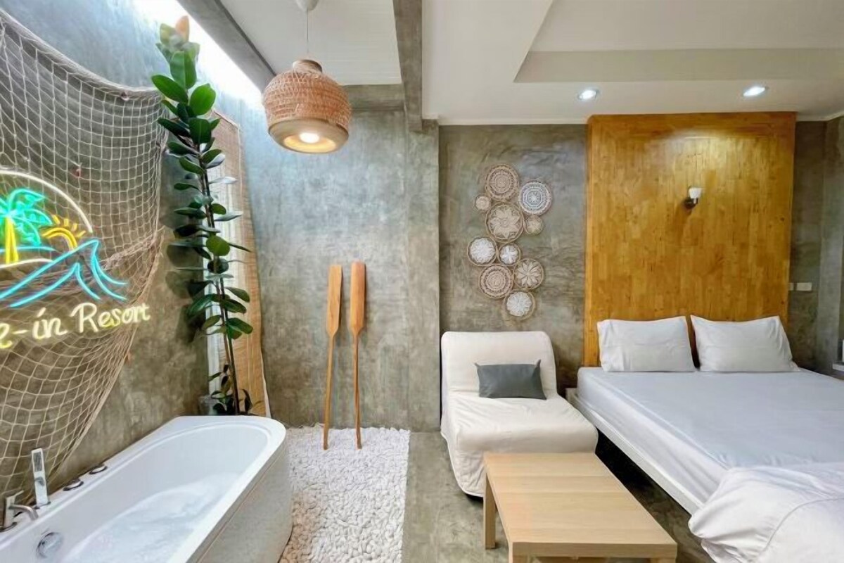 A modern bedroom with a large bed, a small sofa, a wooden headboard, a white bathtub, and a wall with decorative plates.