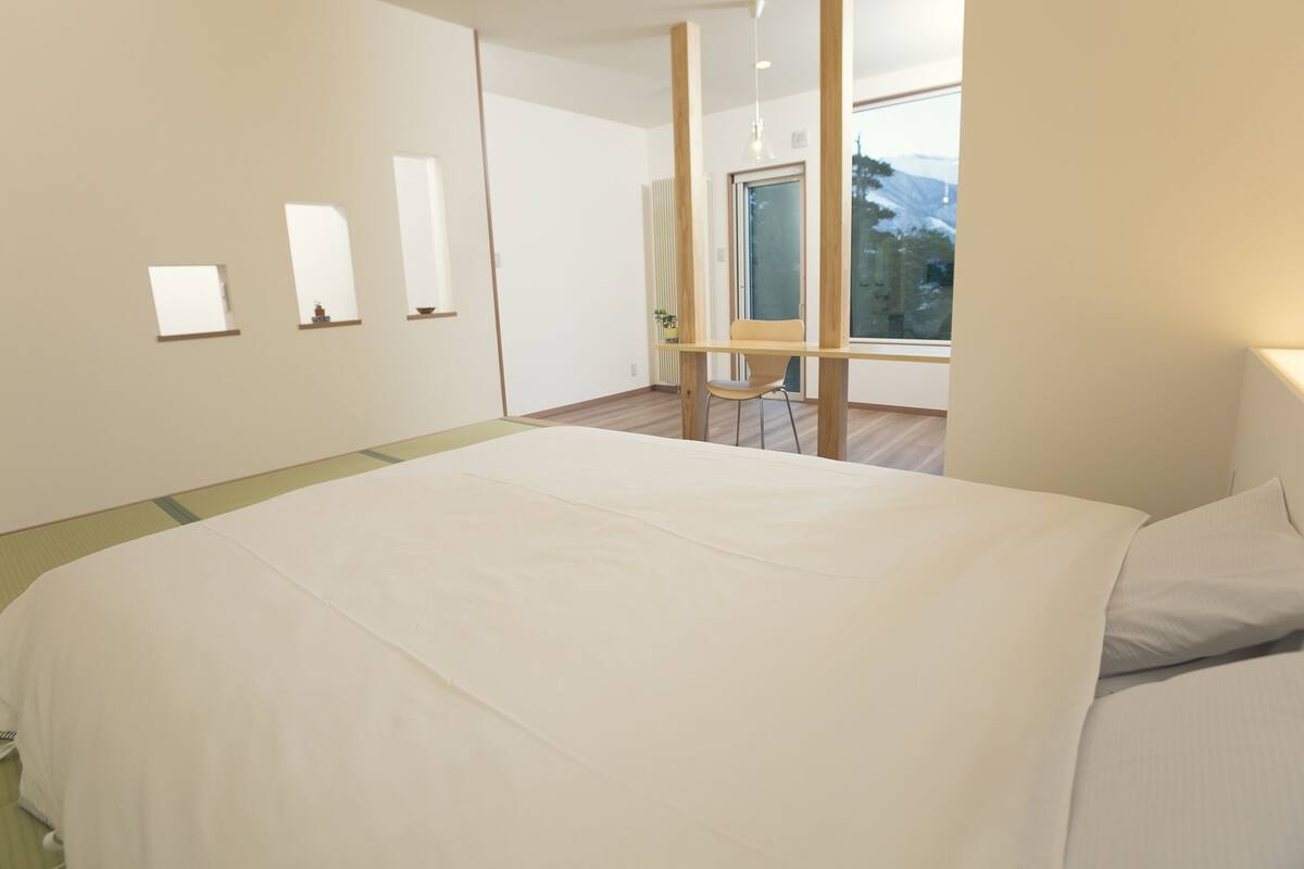Una habitación de hotel moderna con una cama grande, suelo de madera, ventana con vistas y una pequeña mesa con una silla.